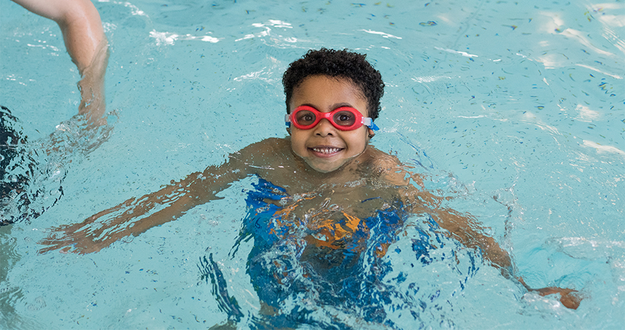 A child in red swimming goggles smiles while swimming at a YMCA health and fitness centre pool.