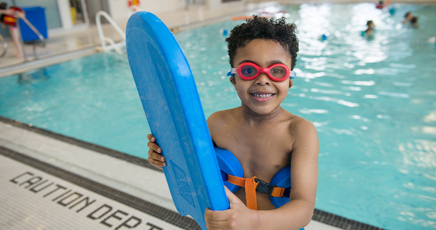 A child in red swimming goggles holds a blue kickboard in front of a YMCA health and fitness centre swimming pool.