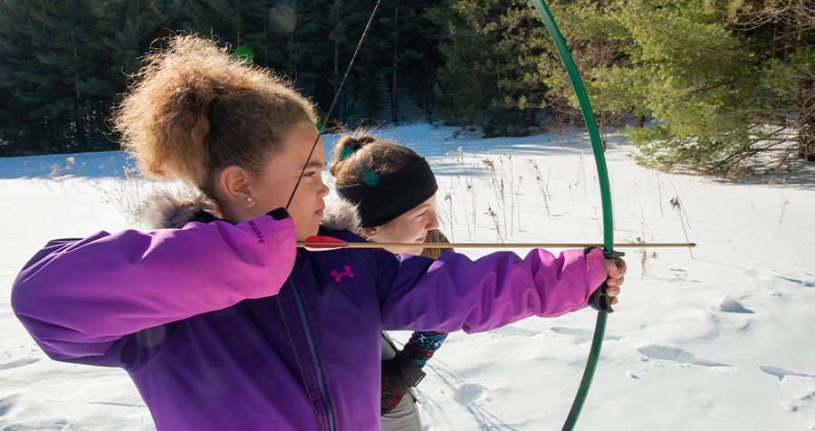 A child in a purple winter coat holds a green archery bow, preparing to shoot at a target, outdoors in the snow at YMCA Camp Pinecrest