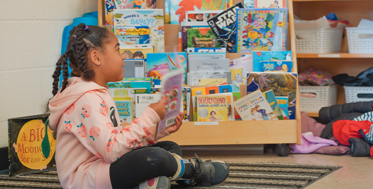 A child sits on a carpet in front of a bookshelf in a YMCA Child Care classroom, reading a picture book.