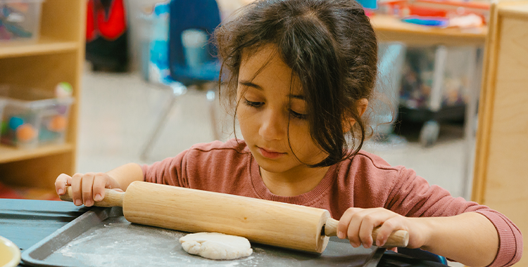 A child uses a rolling pin to roll pizza dough during a cooking activity in a YMCA Child Care classroom.