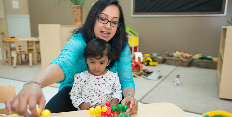 A parent in a bright teal top with glasses and a child in a star patterned top play with building blocks in a YMCA child care classroom
