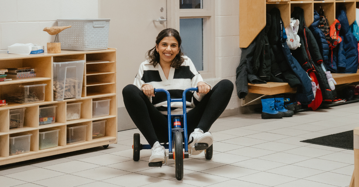 Petra, wearing a white and black striped sweater, sits on a small blue children’s tricycle in a YMCA child care hallway, playfully pretending to ride it.