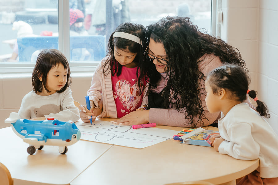 An educator surrounded by three children at a round wooden table guides a child to draw with a blue marker on a sheet of paper. On the table are a box of markers and a blue toy airplane.