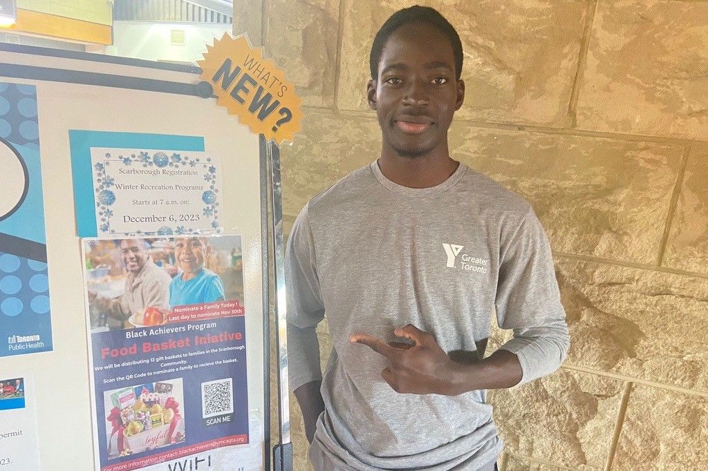 Titobi Oriola advertising for the food drive. He is standing next to a board with a food drive poster. He is wearing a YMCA shirt.