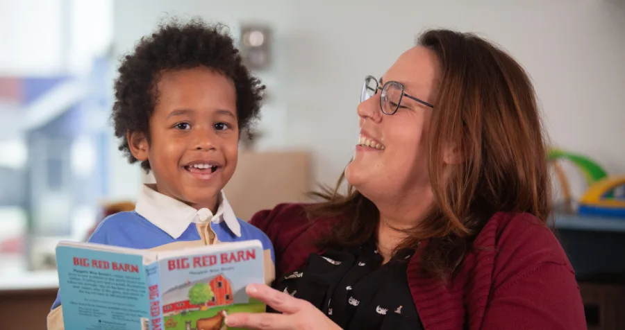 An educator wearing a burgundy cardigan holds the children's book Big Red Barn while reading with a child seated beside them.