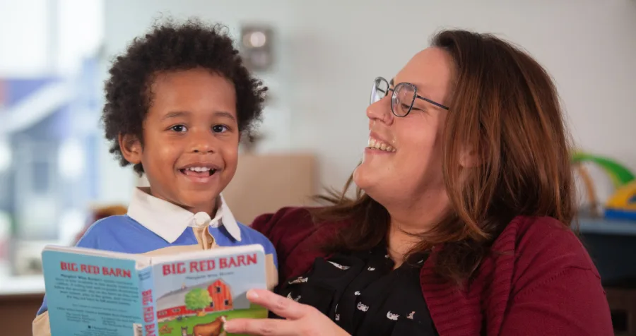 An educator wearing a burgundy cardigan holds the children's book Big Red Barn while reading with a child seated beside them.