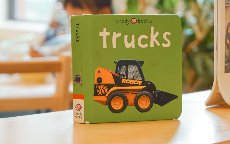 Text: Trucks. Graphic: A green children's board book featuring an orange and black construction vehicle, standing on a wooden table.