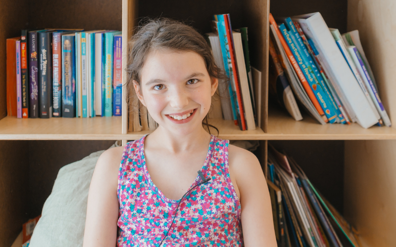 A child wearing a floral dress smiles while seated in a reading area in front of a bookshelf filled with children's books.