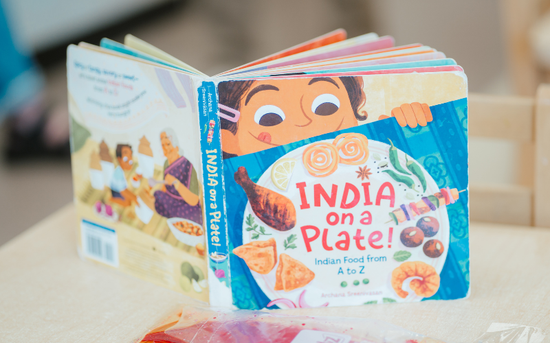 Text: India on a Plate! Indian Food from A to Z. Graphic: A colourful children's board book featuring illustrations of Indian dishes and a child peeking over a plate of food.