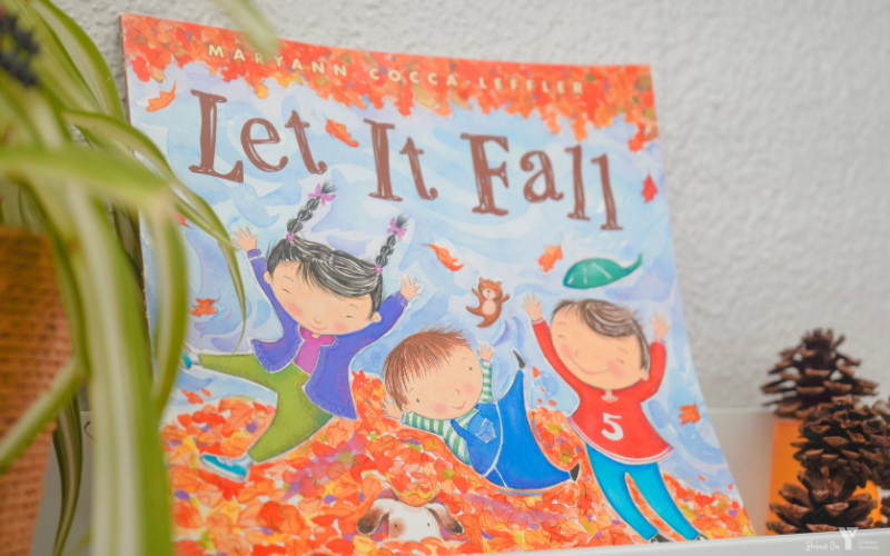 Text: Let It Fall. Graphic: A children's book cover featuring three illustrated children playing in a pile of colourful autumn leaves, with falling leaves and a squirrel around them.