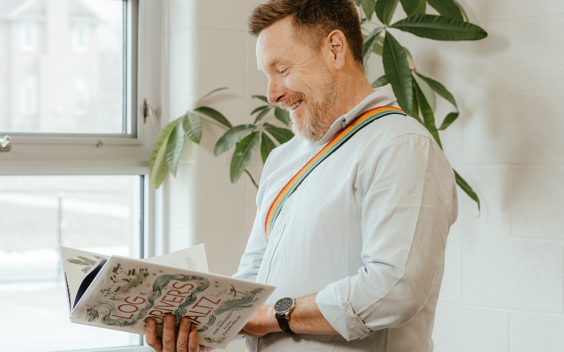 An adult wearing a white shirt and rainbow-coloured strap holds an open copy of The Log Driver’s Waltz by Wade Hemsworth, illustrated by Jennifer Phelan, beside a window.