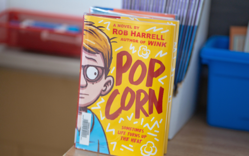 Text: Popcorn by Rob Harrell. Graphic: A yellow book cover featuring an illustrated boy with glasses and red lettering, displayed on a shelf.
