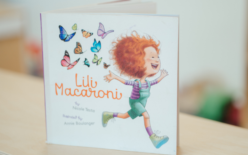 Text: Lili Macaroni. Graphic: A children's book cover featuring a smiling, red-haired girl running with her arms outstretched, surrounded by colourful butterflies.