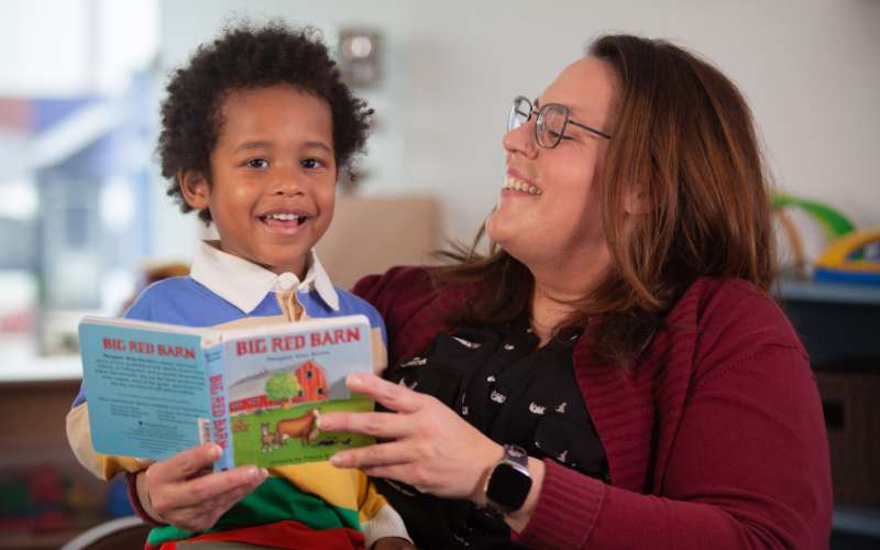 An educator wearing a burgundy cardigan holds the children's book Big Red Barn while reading with a child seated beside them.