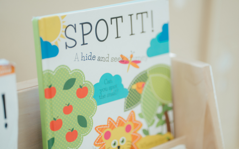 Text: Spot It! A hide and seek. Graphic: A colourful children's book featuring illustrated trees, apples, a dragonfly and a lion.