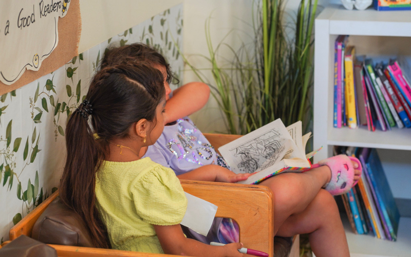 Two children sit together on a wooden bench in a reading area, looking at an open illustrated book, with a bookshelf and plants nearby.