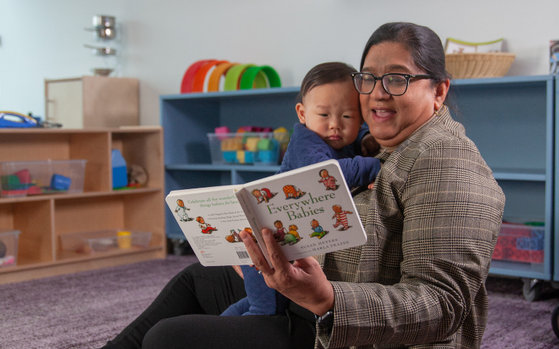 An educator wearing a plaid shirt holds an open copy of Everywhere Babies by Susan Meyers, illustrated by Marla Frazee, while seated in a child care setting.