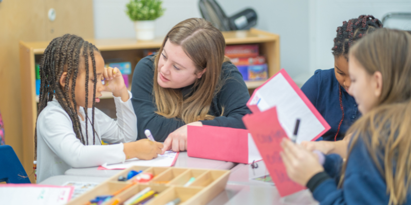 A child care educator comforts a child working on a writing activity at a table with two other children.