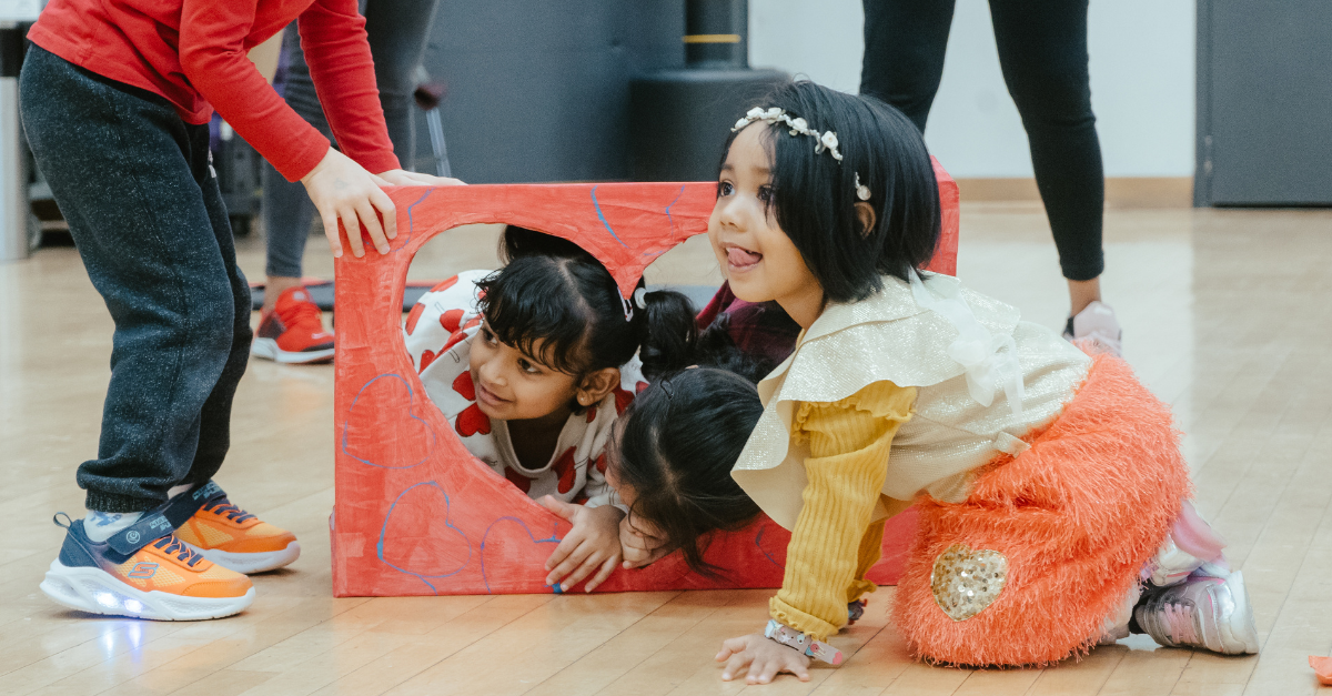 Three children crawl through a red heart-shaped cardboard cutout as part of a physical activity obstacle course.