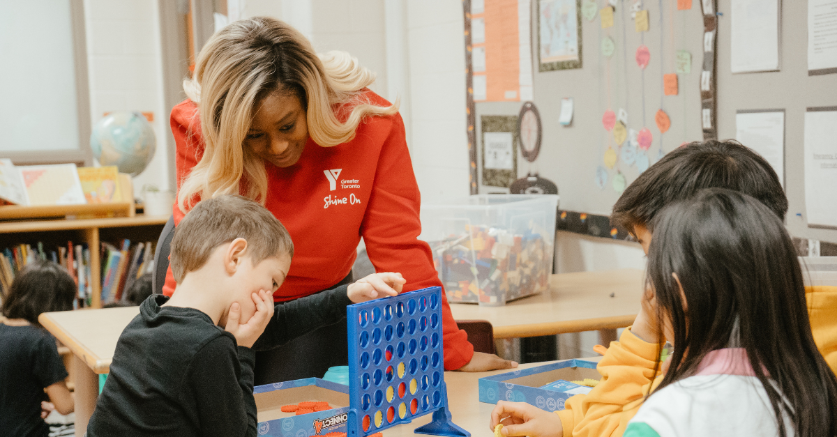Susan, wearing a red YMCA of Greater Toronto Shine On logo sweater, watches children play Connect Four in a before and after school classroom.