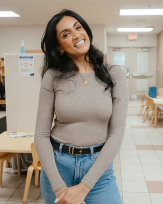 Vanessa, wearing a light brown long-sleeve top and blue jeans, smiles while standing in a YMCA child care hallway.