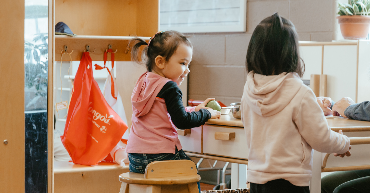 Two children explore a play kitchen together at a YMCA child care classroom, wearing pink and light-pink hoodies.