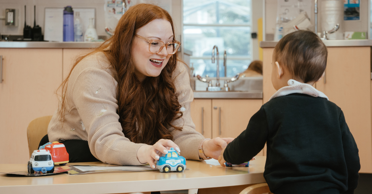 Alyssa, wearing a beige long-sleeve top, plays with toy cars alongside a toddler in a black sweater in a YMCA toddler classroom.