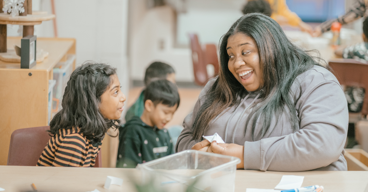 Shianne engages with a school-age child wearing a striped long-sleeve shirt as they play a paper fortune teller game together in a YMCA program space.