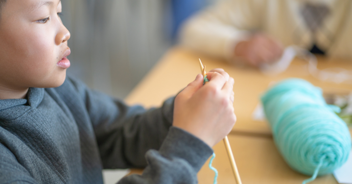 A child in a charcoal gray hoodie holds crochet needles at a table in a Before & After School program, with teal yarn in the background.