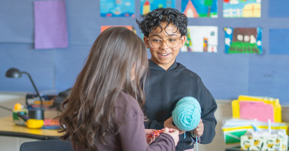 A child wearing glasses and a black hoodie speaks with another child while holding a ball of blue teal yarn.