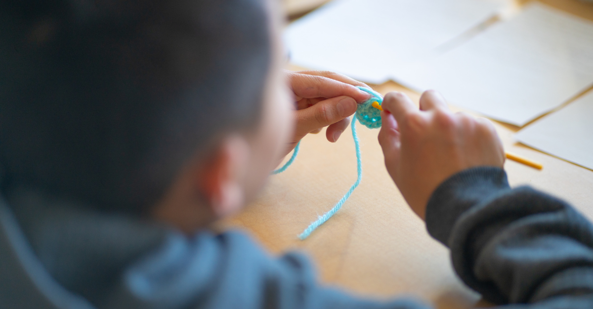 Close-up of a child’s hands in a charcoal hoodie using a yellow crochet needle to thread blue yarn at the start of a project.
