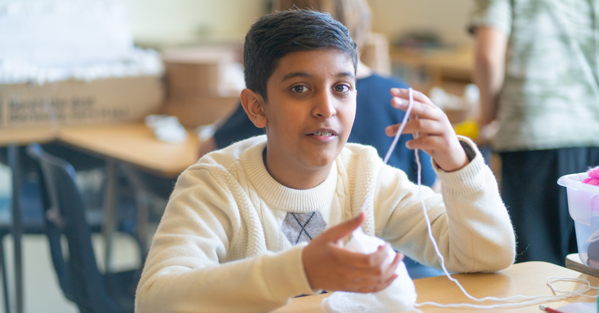 A child in a beige sweater smiles while working with white thread on a crochet project.