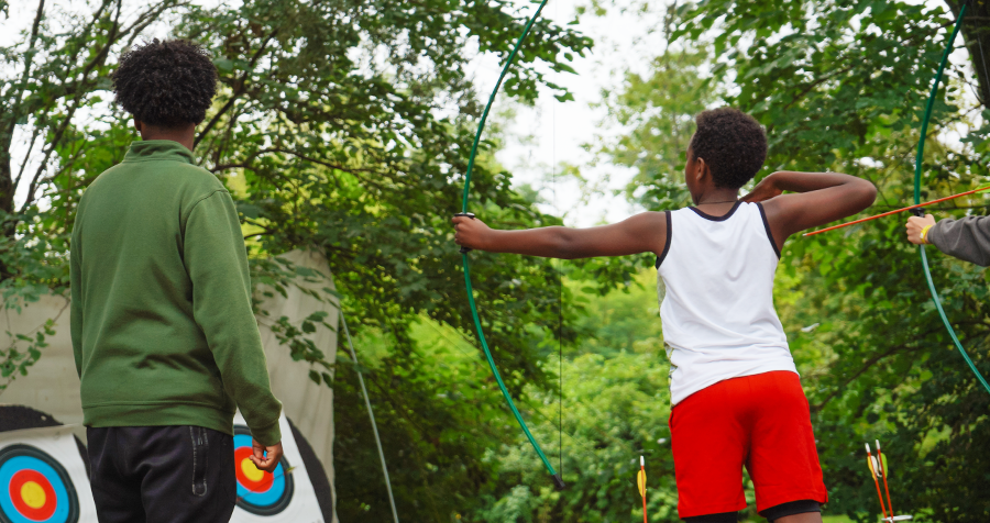  child in a white tank top and red shorts aims an arrow at a target on lush green YMCA campgrounds, accompanied by a camp counselor wearing a green sweater.