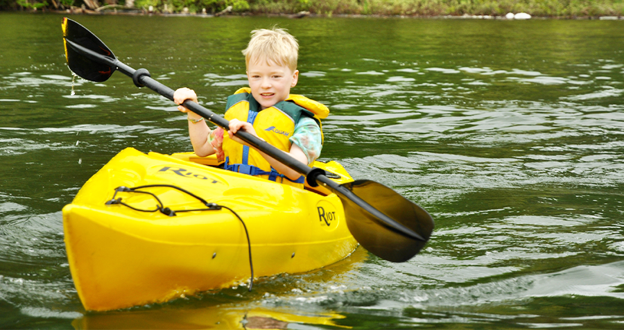 A child, adorned in a yellow lifejacket, paddles a yellow kayak during a YMCA summer camp program