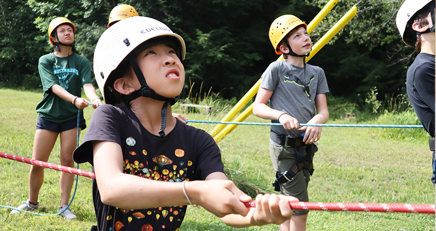 Four determined children, dressed in helmets, graphic tees, and rock climbing gear, exert effort as they pull on a rope to assist a fellow camper in climbing at the YMCA's Camp Cedar Glen program.