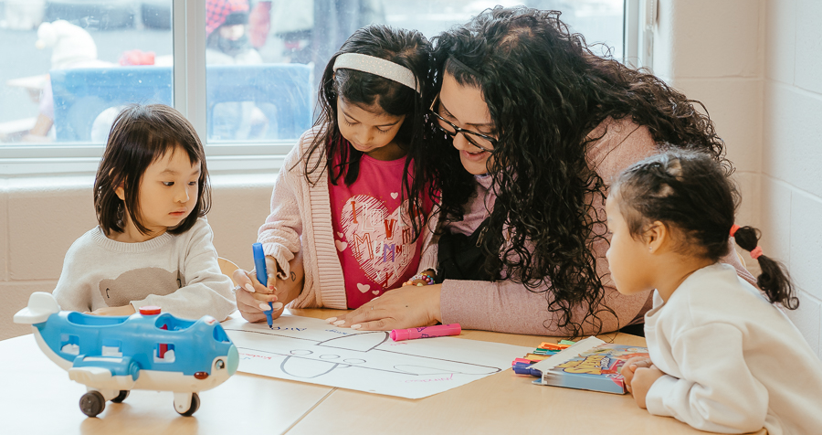 Josie helps a child in a bright pink t-shirt and light-pink cardigan use a purple marker to draw a spaceship on paper. Two children observe.