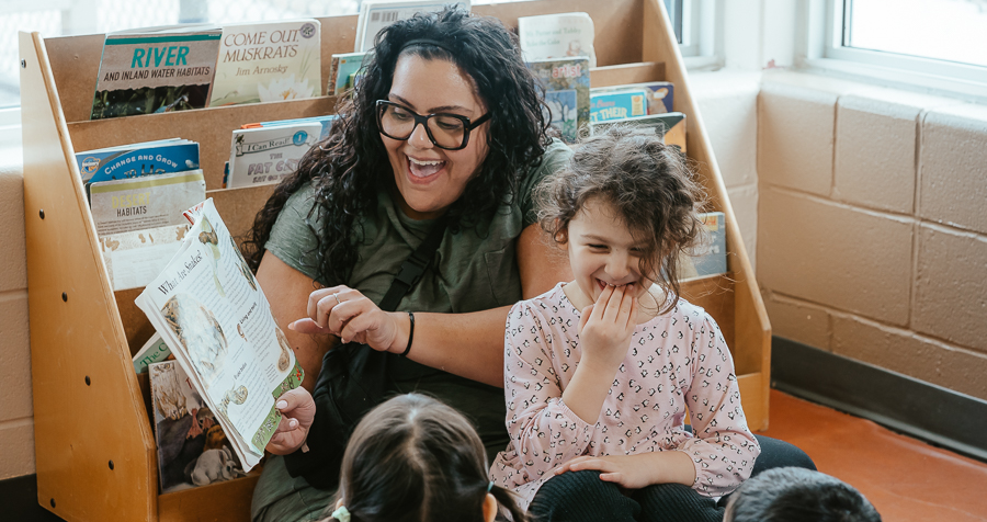 Sitting in the reading nook of a YMCA child care classroom, Josie reads a picture book to the children. A child with brown hair wearing a pink dress laughs while sitting in Josie's lap.