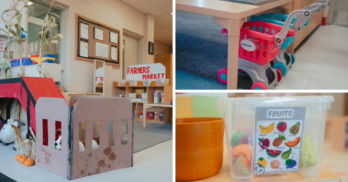 Fall-themed dramatic play area in a classroom with a cardboard barn, toy animals, and mini pumpkins. Nearby are toy shopping carts, a "Farmers Market" sign on shelves, and a plastic bin filled with play food labeled "Fruits."