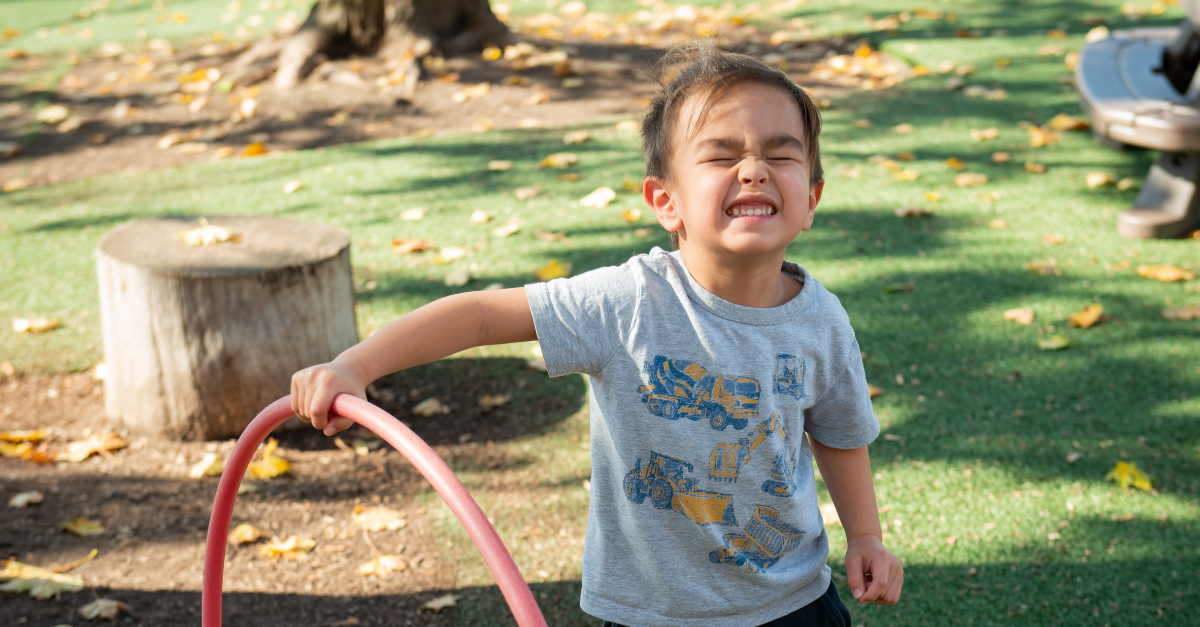 A smiling child wearing a grey top with truck designs holds a red hula hoop outdoors on a Y M C A child care playground in the fall. Yellow leaves cover the grass around them.