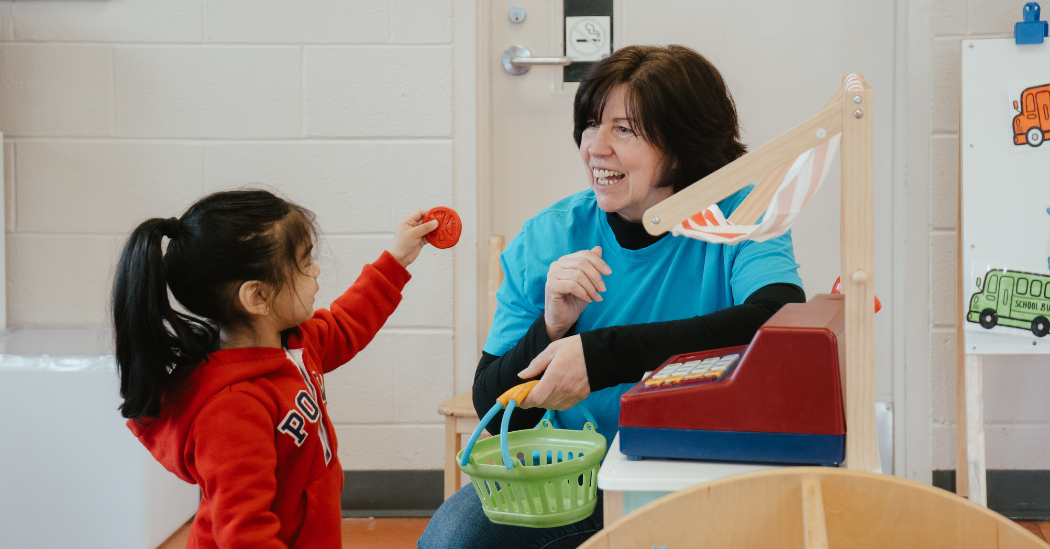 Valerie, wearing a bright blue YMCA shirt and black long-sleeve top, holds a green toy grocery basket beside a toy cashier. A child in a red hoodie offers a toy tomato slice as Valerie engages with them in a YMCA child care classroom.
