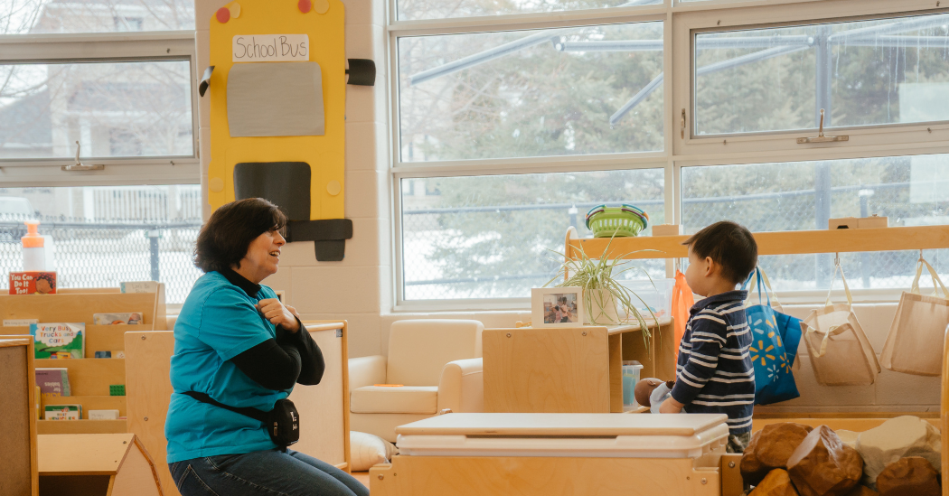 Valerie kneels on the floor with arms crossed in a positive X gesture across their chest, wearing a bright blue YMCA shirt over a black long-sleeve top. They sing a children’s song while a child in a navy striped shirt holding a baby doll looks toward them in a YMCA child care classroom.