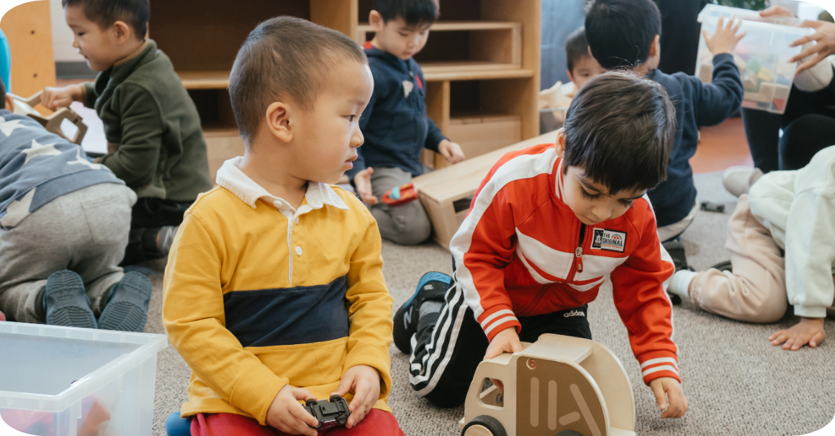 Two children playing with toy cars and wooden vehicles on a carpet while other children play in a busy YMCA preschool room.