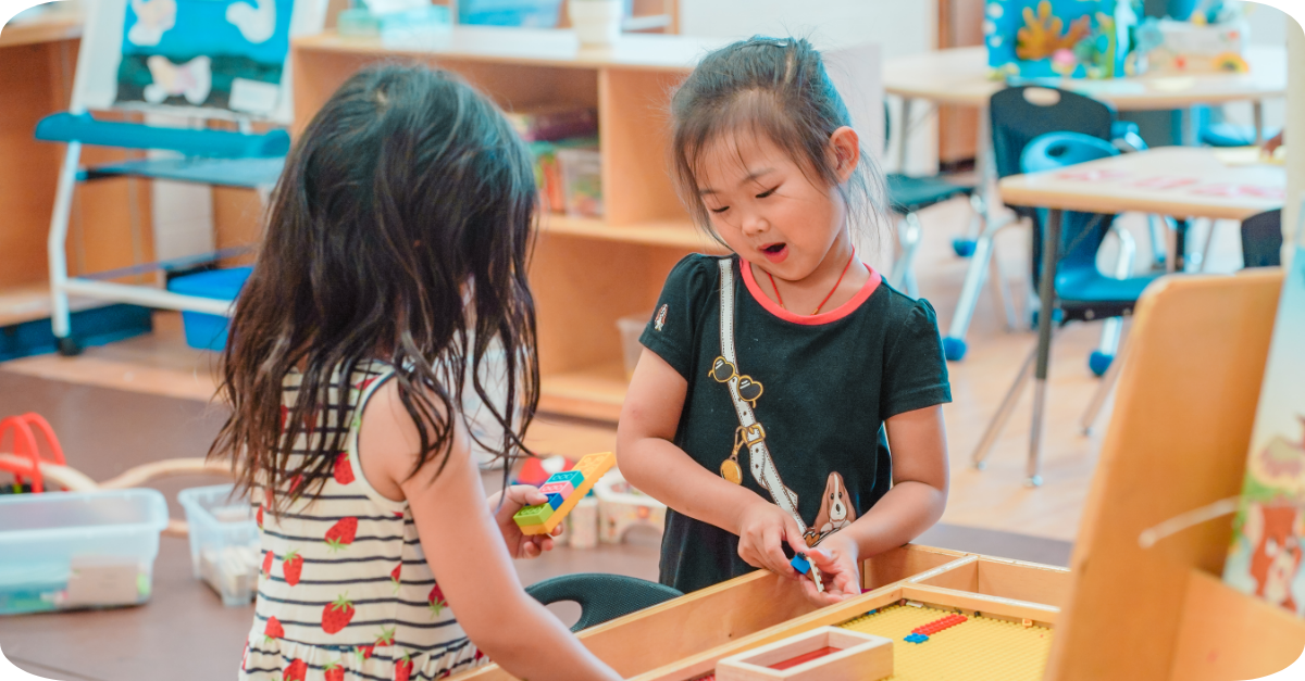 Two children standing at a table sharing Lego pieces and building together in a YMCA room.