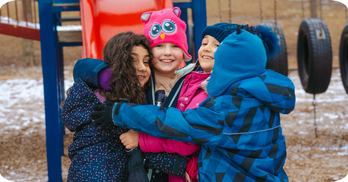 Four school-age children in winter clothing hugging together on an outdoor playground.