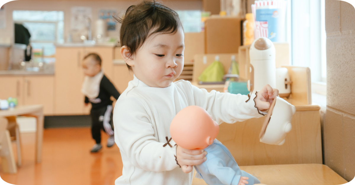 Child engaging in independent play while pretending to feed a toy baby with a toy pot in a YMCA room.