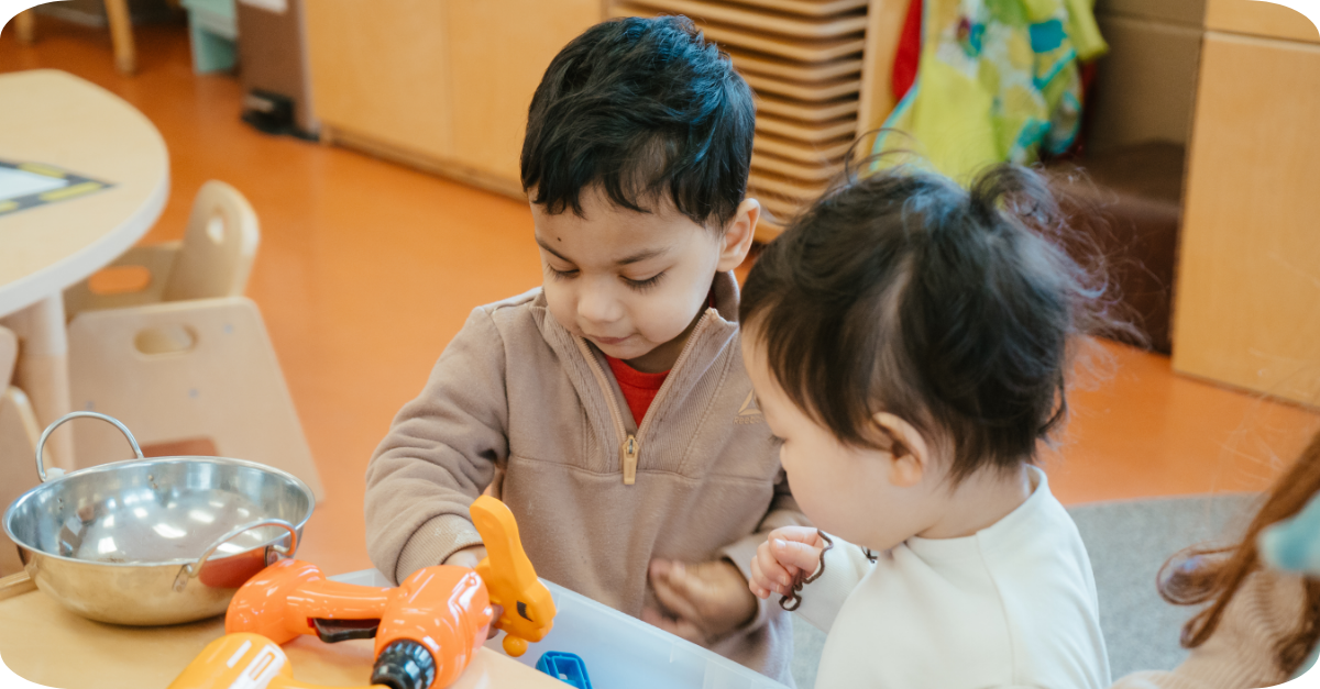 Two children exploring a drawer of toys beside a low table with a bowl and toy drill in a YMCA toddler room.