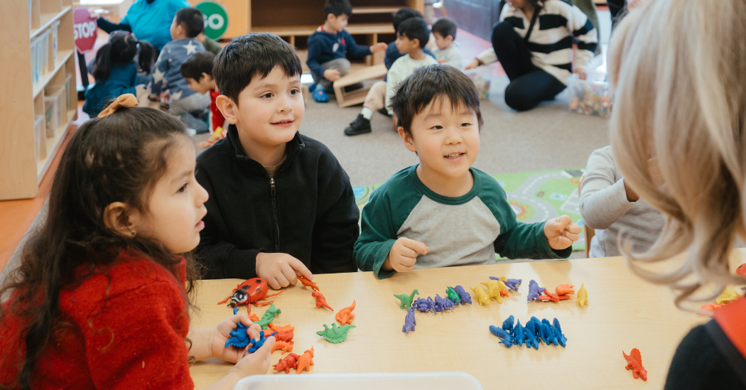 Three children smile as they play with colourful toy dinosaurs at a table, engaging with an educator in a busy preschool classroom.