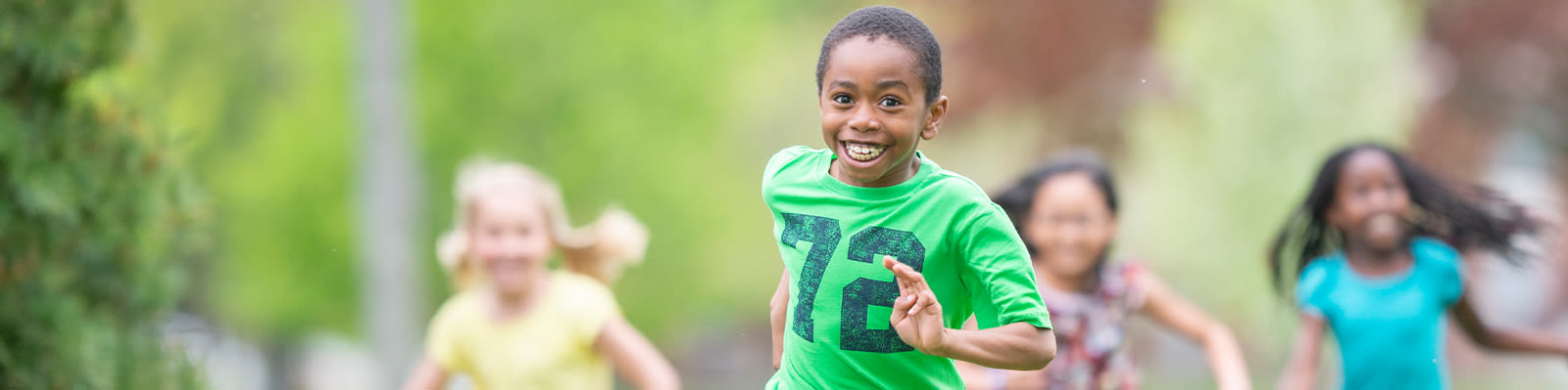 A smiling child in a green shirt runs towards the camera. Three children in colourful shirts run behind them.
