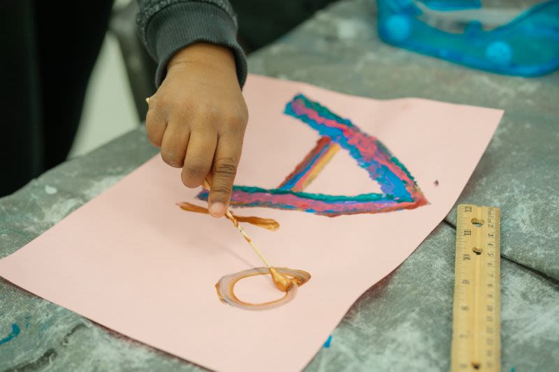 A child’s hand holds a paint brush while painting on pink construction paper. The artwork features colourful, raised shapes outlined in blue, pink, and gold. A wooden ruler and a blue tray are visible on the table nearby.